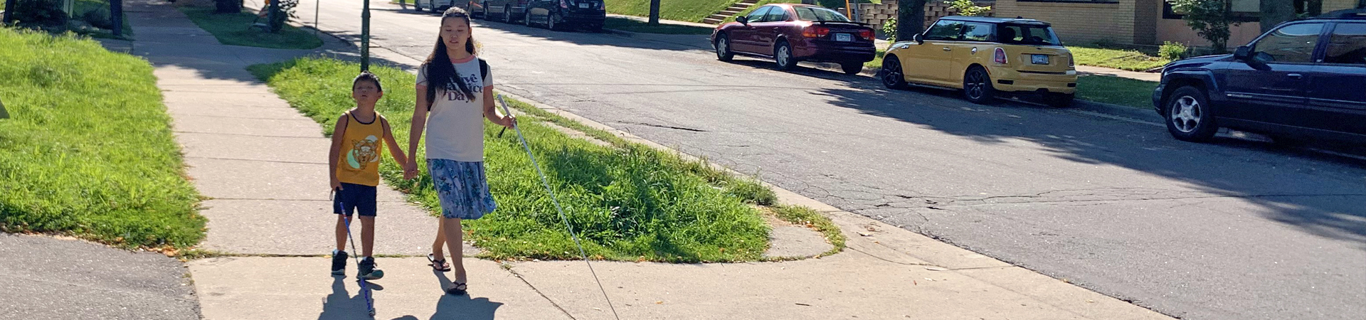 A young, blind person walks down the sidewalk with his cane and blind mentor.